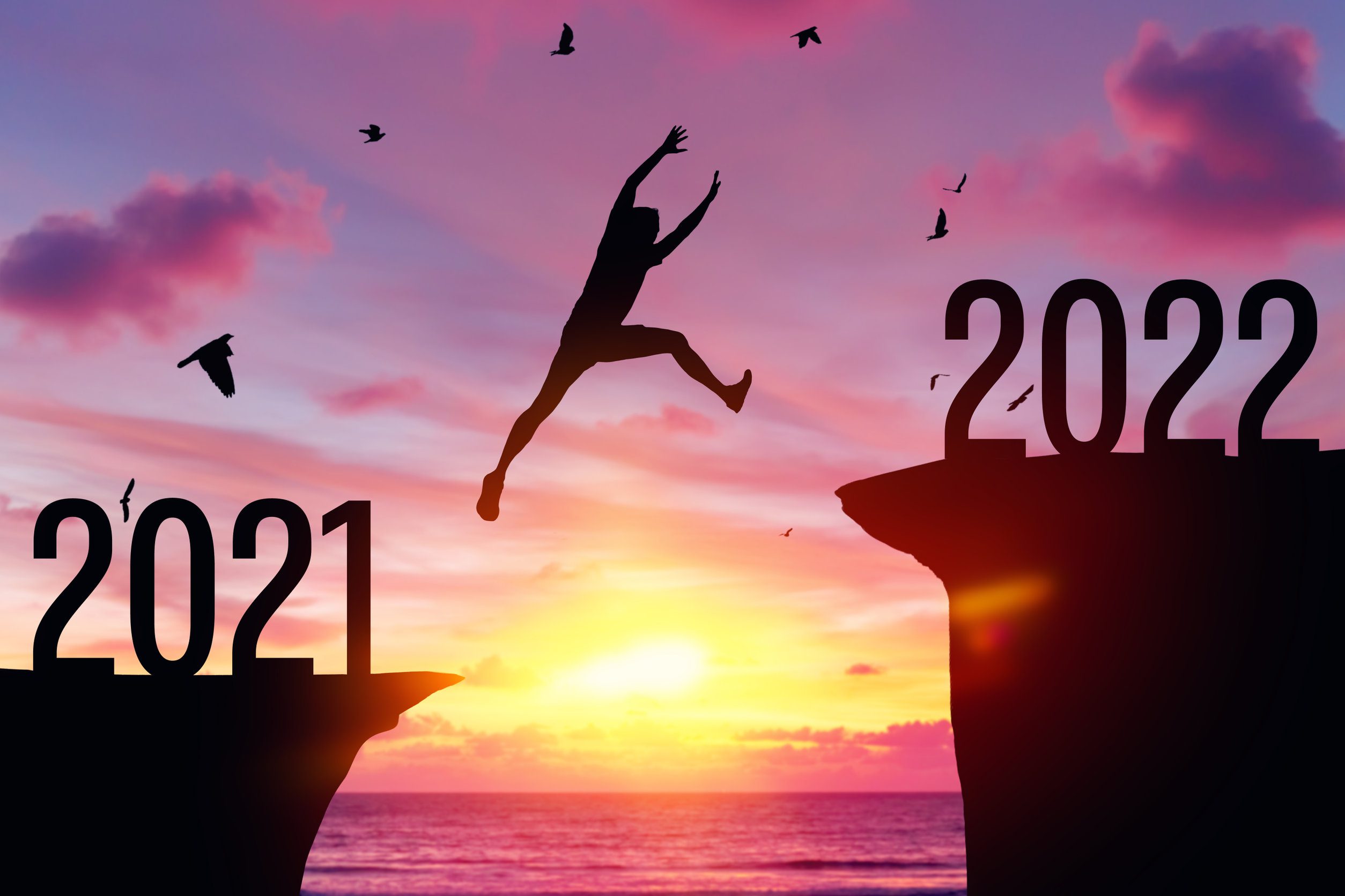 Silhouette man jumping between cliff with number 2021 to 2022 and birds flying at tropical sunset beach. 2022 عام تصفية الحسابات والتمادي في الطغيان